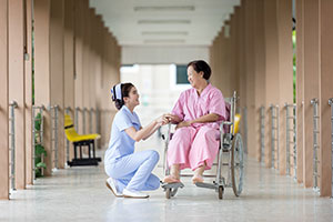 Nurse Kneeling, Talking To Female Patient In A Wheelchair In The Hallway.