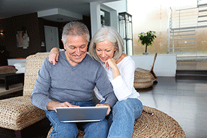 A Senior Couple Using A Computer At Home, Smiling While Looking At The Screen.