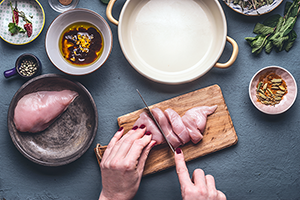 Close-up Of Woman Hands Cutting Chicken Breast On A Cutting Board, Surrounded With Food And Cookware.