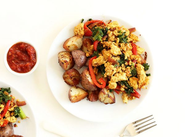 Heart-healthy breakfast: tofu scrambled with kale, red pepper, baked potatoes on white plate besides chili garlic cumin sauce.