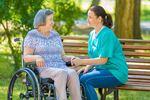 Personal Support Worker On Park Bench Holding Hands With Senior Woman On Wheelchair.