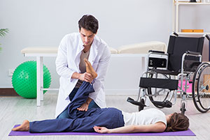 Male Physiotherapist Pressing Patient's Leg Down, Patient Lying On Mat In A Physiotherapy Clinic.